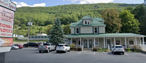 A two-story building with a green roof, surrounded by trees and parked cars, set against a mountainous backdrop.