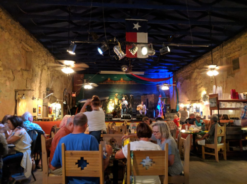 A lively restaurant scene with diners enjoying food while a band performs on stage, decorated with Texas flags.