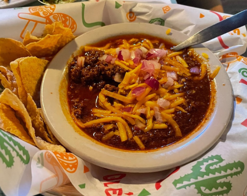 A bowl of chili topped with cheese and onions, served with tortilla chips on the side.