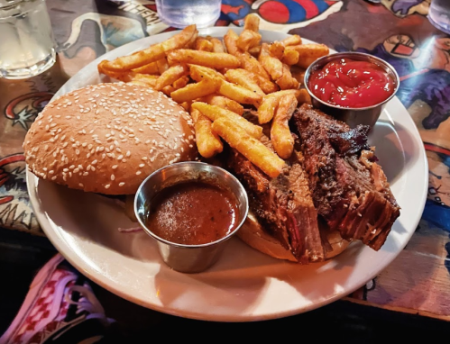 A plate with a sesame seed bun, brisket, fries, and two dipping sauces on a colorful table.