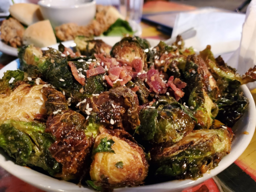 A bowl of crispy Brussels sprouts topped with bacon bits and sesame seeds, served in a casual dining setting.