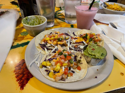 A plate of tacos topped with cabbage, mango, and salsa, served with guacamole and drinks in the background.