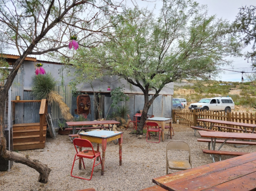 Outdoor seating area with rustic tables, a tree, and a decorative lion head on a wall in a desert setting.