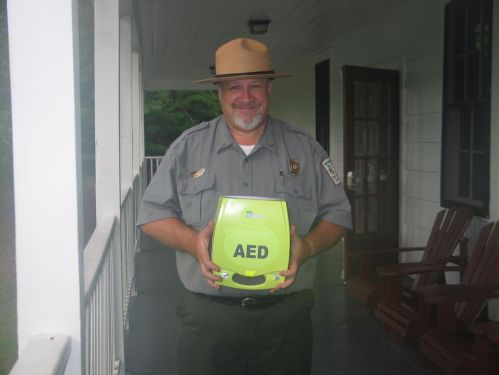 A park ranger smiles while holding a bright green AED device on a porch.