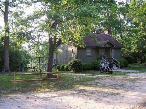 A small green house surrounded by trees, with a motorcycle parked nearby and a picnic area in the background.
