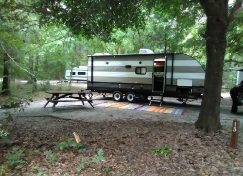 A camper parked in a wooded area, with a picnic table and colorful rug nearby, surrounded by trees and leaves.
