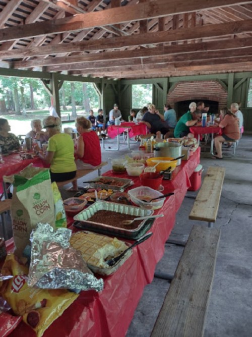 A picnic shelter filled with people enjoying a potluck meal, with tables covered in food and red tablecloths.