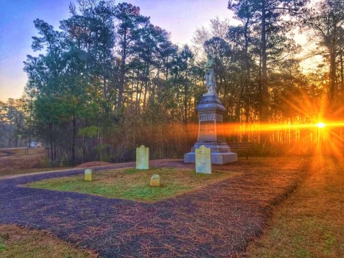 A serene cemetery at sunrise, featuring a monument surrounded by trees and gravestones, with sunlight streaming through.