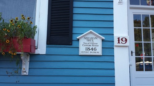 Sign on a blue building reads "Abraham True, Naval Officer, Custom House, 1846, Historic Salem, Inc." with flowers nearby.
