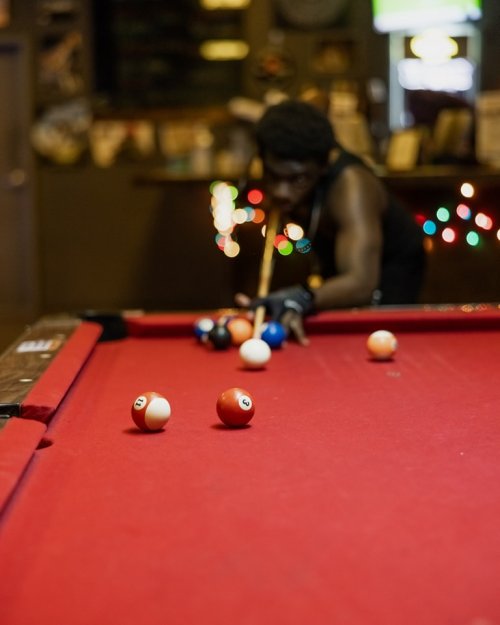 A person playing pool, focusing on a shot, with colorful balls on a red felt table and festive lights in the background.