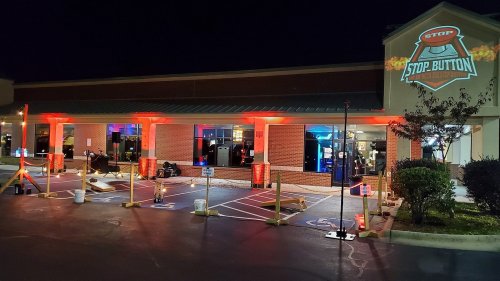 A brightly lit storefront at night with red lights and a "Stop Button" sign, featuring outdoor setup and parking spaces.