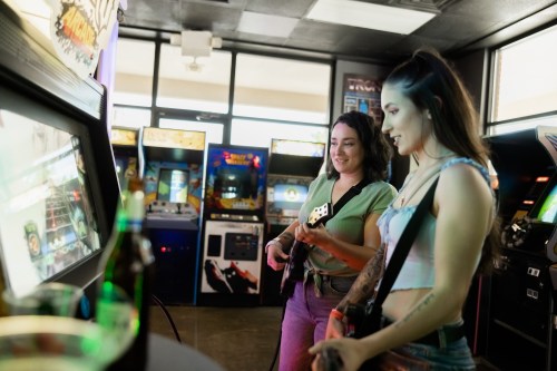 Two women play a video game in an arcade, smiling and holding controllers, with arcade machines in the background.