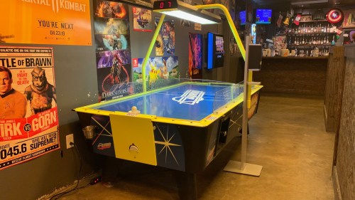 A brightly colored air hockey table in a game room with posters on the walls and a bar in the background.