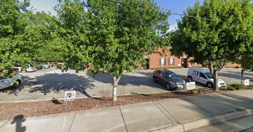 A parking lot with several cars and trees, adjacent to a brick building. Signs are visible in the foreground.