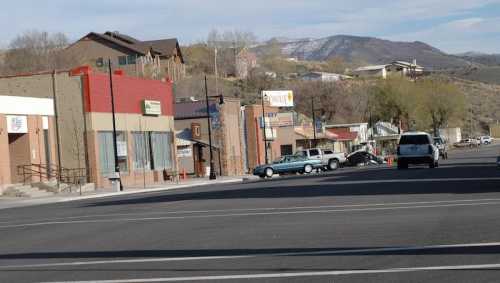 A small town street with shops, parked cars, and mountains in the background under a clear blue sky.