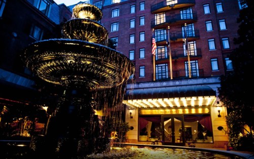A lit fountain in front of a hotel with elegant architecture and warm lighting at dusk.