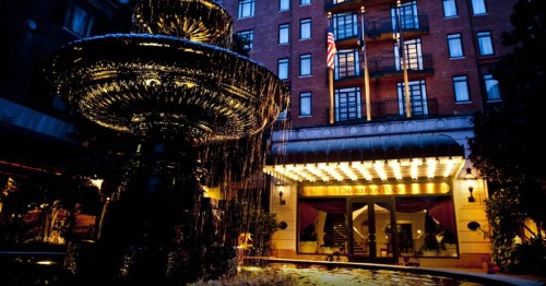 A lit fountain in the foreground with a hotel entrance and American flag in the background, illuminated at night.