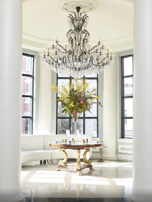 Elegant room featuring a large chandelier, a floral centerpiece on a round table, and tall windows with natural light.