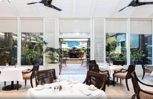 Bright restaurant interior with white decor, tables set for dining, and a view of a lush garden and fountain outside.
