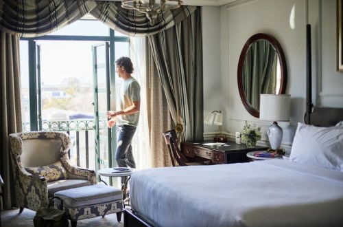 A man stands by an open window in a stylish hotel room, looking outside. Elegant furnishings are visible.