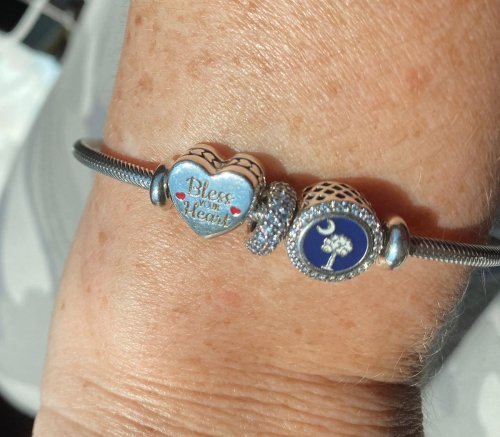 A close-up of a wrist adorned with a charm bracelet featuring a heart charm that says "Bless your Heart."