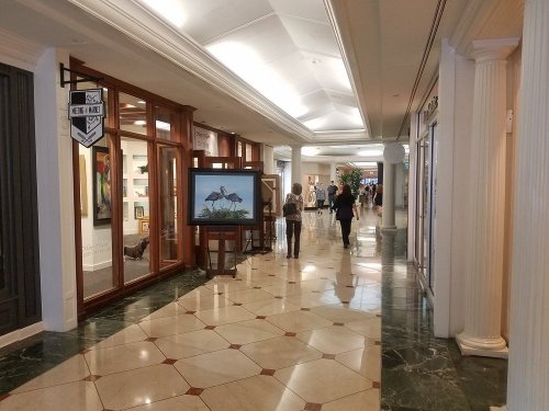 A spacious indoor corridor with shops, featuring a large screen displaying artwork and people walking by.