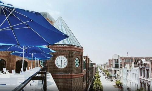 A view of a city street with a clock tower and blue umbrellas on a balcony, under a clear sky.