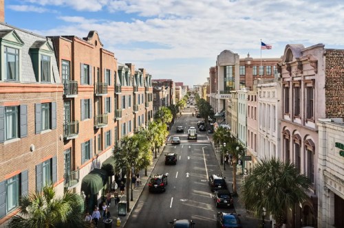 A bustling city street lined with historic buildings, palm trees, and cars under a partly cloudy sky.