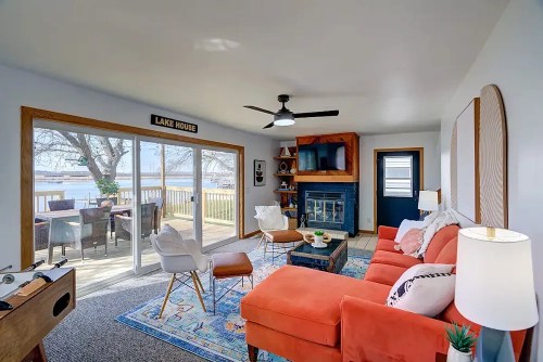 Cozy living room with orange sofa, fireplace, and large windows overlooking a deck and lake. Bright and inviting atmosphere.