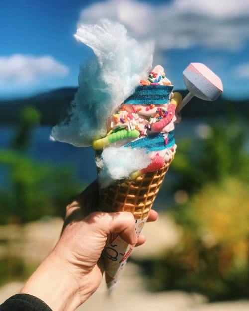 A colorful ice cream cone topped with cotton candy, sprinkles, and a marshmallow, held against a scenic background.