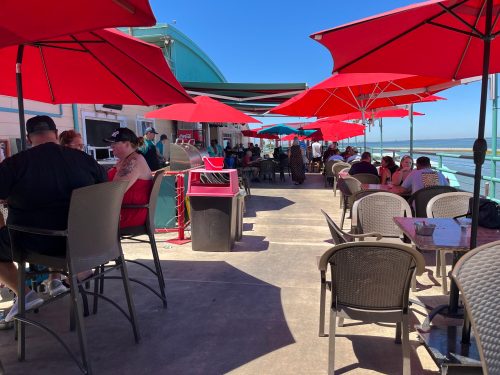 Outdoor dining area with red umbrellas, people seated at tables, and a view of the water in the background.