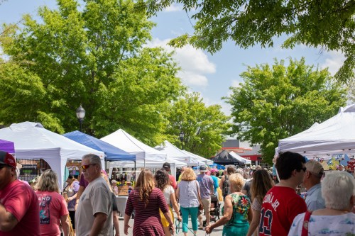 A bustling outdoor market with people browsing tents under green trees and a blue sky.