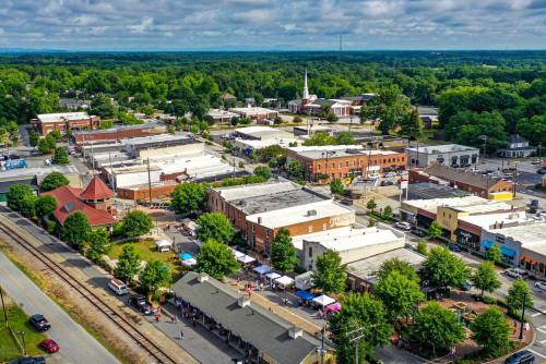 Aerial view of a small town with shops, trees, and a train station, surrounded by green hills under a cloudy sky.