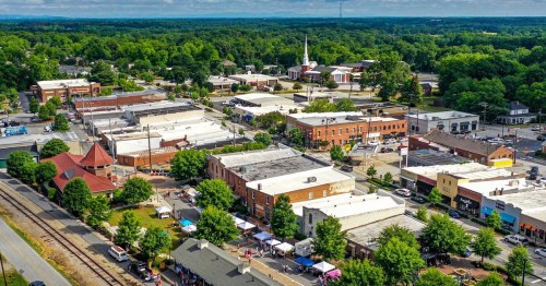 Aerial view of a small town with shops, trees, and a church, surrounded by greenery and a clear blue sky.