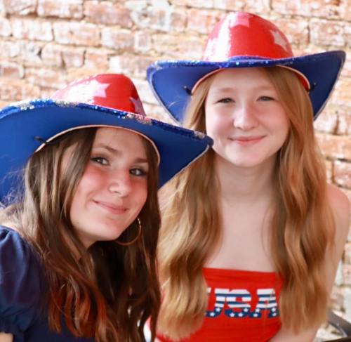 Two smiling girls wearing blue and red cowboy hats, posing together against a brick wall.