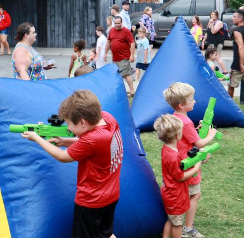 Children in red shirts play with toy guns behind large blue inflatable barriers at a lively outdoor event.