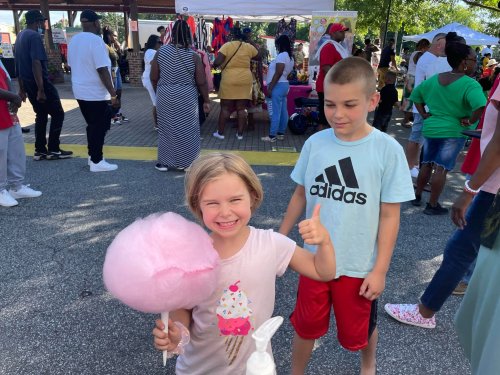 A young girl smiles and gives a thumbs up while holding pink cotton candy at a lively outdoor event.