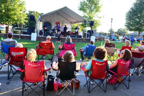 A crowd sits in lawn chairs enjoying a live band performance under a tent in a park. Trees and a stage are visible.