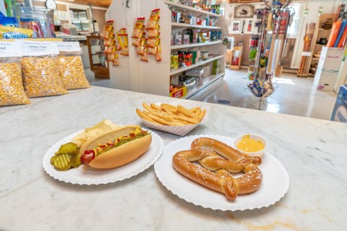 A hot dog with pickles, a pretzel with cheese, and fries on white plates, set against a store interior.