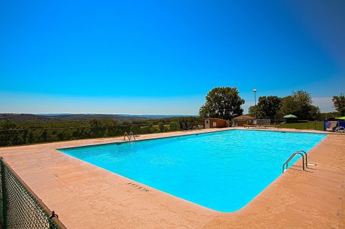 A clear blue swimming pool surrounded by green trees and hills under a bright blue sky.