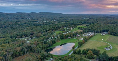 Aerial view of a lush green landscape with a pond, surrounded by trees and rolling hills under a colorful sky.