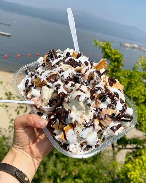 A hand holding a large dessert bowl filled with ice cream, chocolate sauce, and toppings, with a scenic waterfront in the background.