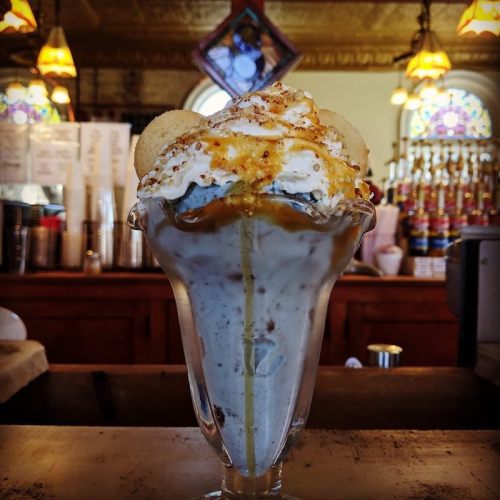 A tall glass of ice cream topped with whipped cream, caramel, and cookies, set against a vintage café backdrop.