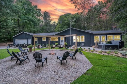 A cozy black house surrounded by trees, with a fire pit and seating area in the foreground under a colorful sunset.