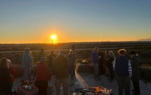 A group of people gather outdoors at sunset, enjoying food and drinks with a scenic landscape in the background.