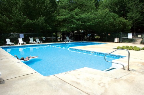 A clear blue swimming pool surrounded by lounge chairs and greenery, with a person relaxing on a float.