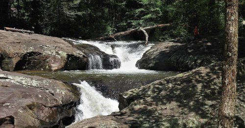 A serene waterfall cascades over rocky terrain, surrounded by lush green trees and a person in the distance.