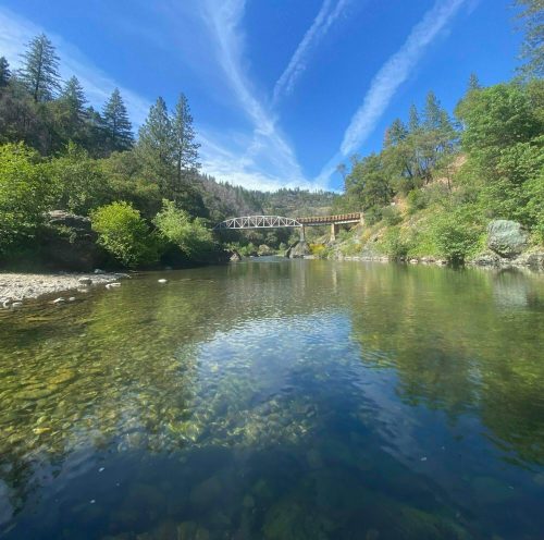 A serene river scene with clear water, lush greenery, and a bridge under a blue sky with wispy clouds.