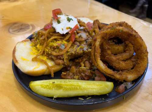A plate of food featuring a burger with toppings, onion rings, a pickle, and a side of chili with beans.