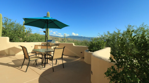 A patio with a table and chairs under a green umbrella, surrounded by desert plants and mountains in the background.
